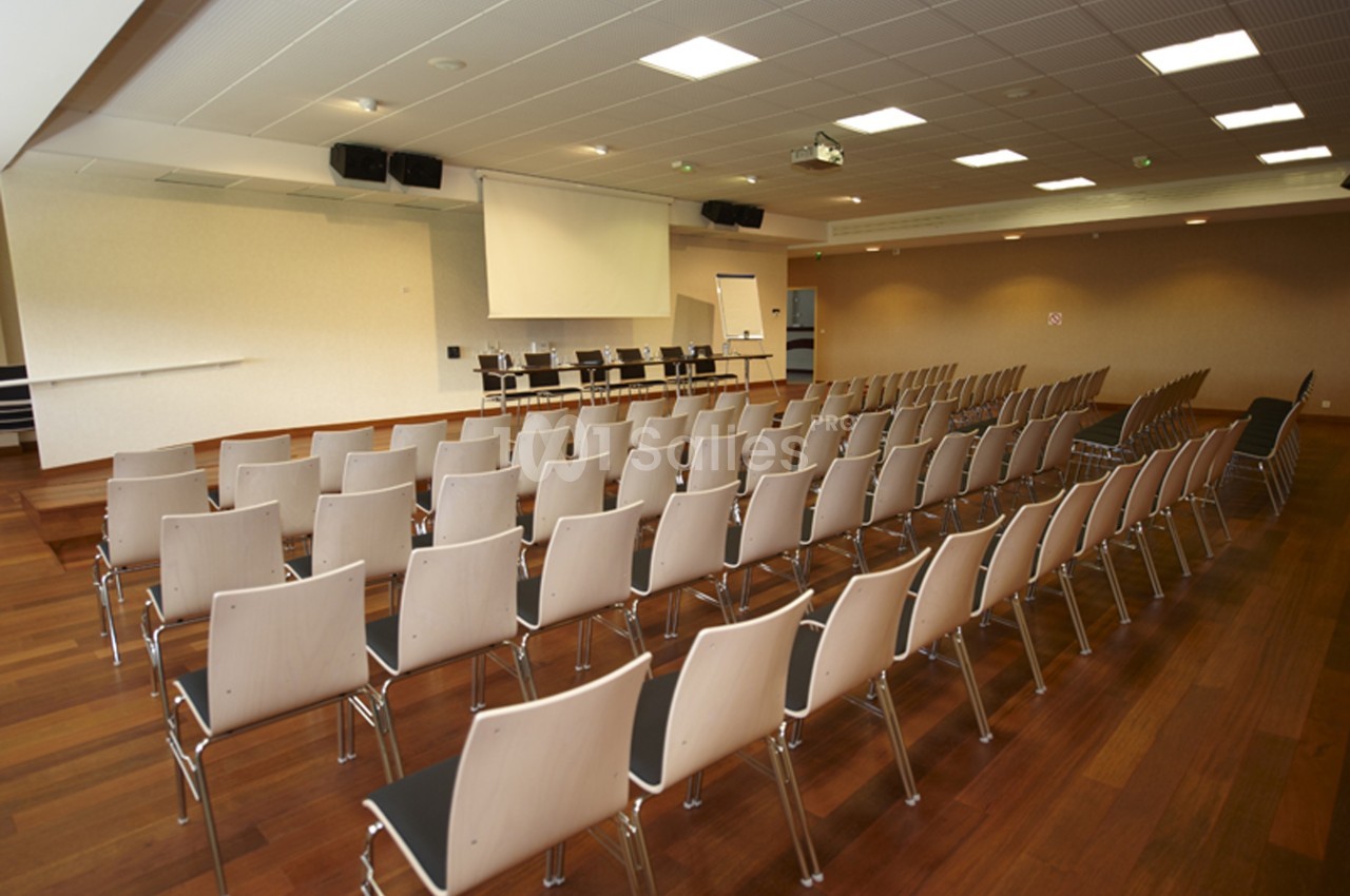 Salle de conférence vide avec rangées de chaises alignées, écran de projection et tableau blanc à l'avant.