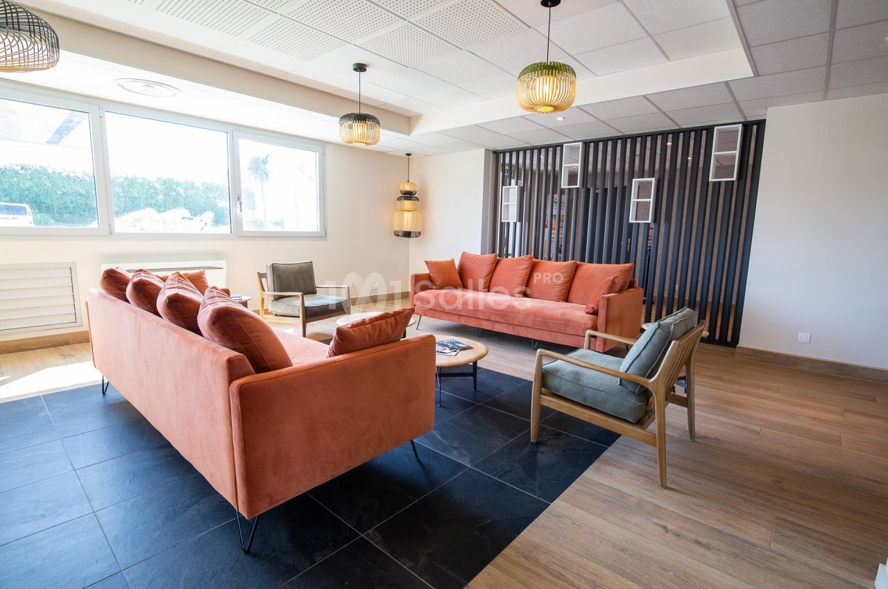 Salon moderne avec canapés orange, fauteuils en bois, sol en carrelage et parquet, et grande fenêtre lumineuse.