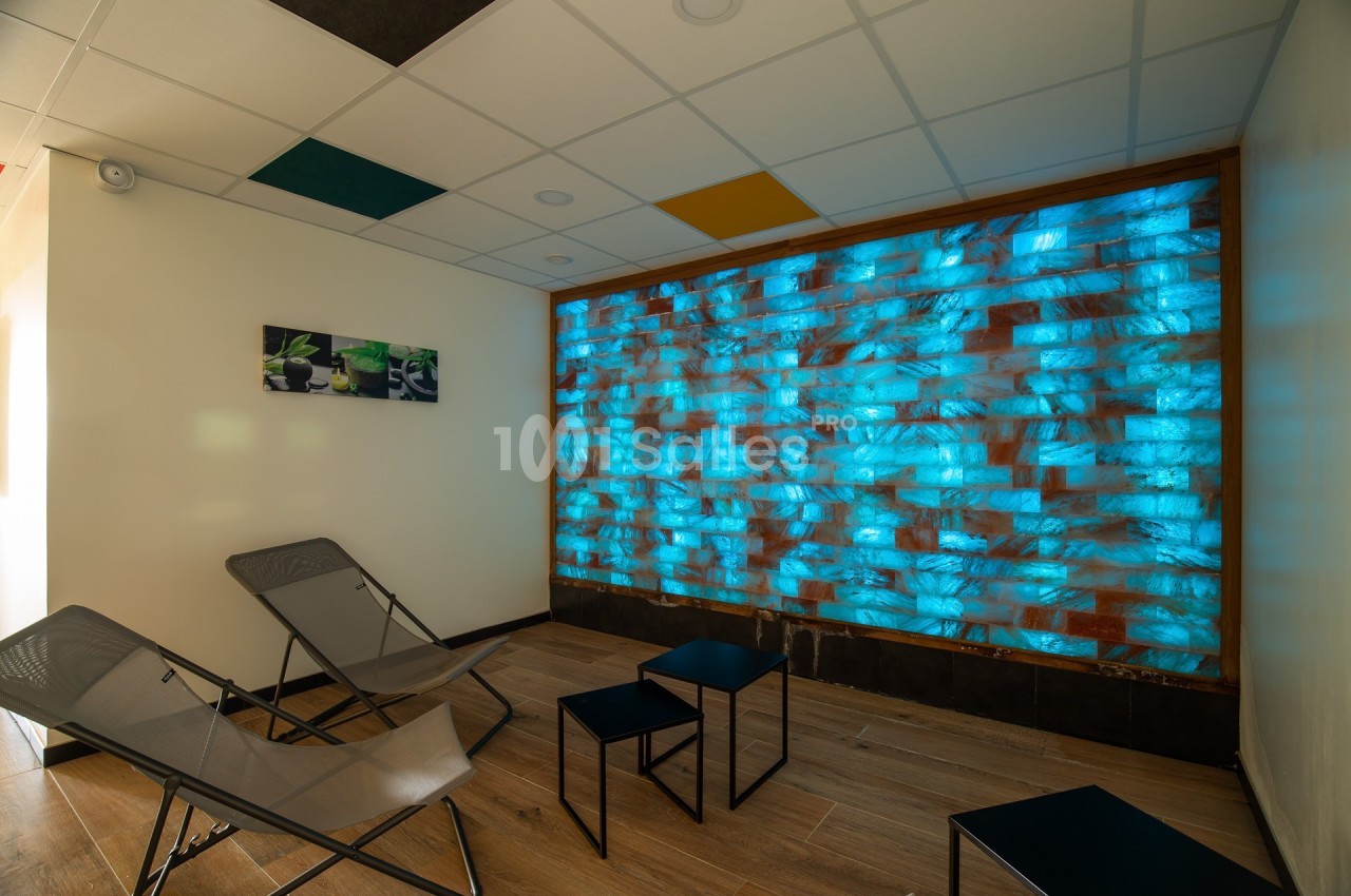 Mur lumineux bleu dans une salle de détente avec chaises longues, tables basses et plafond à dalles colorées.