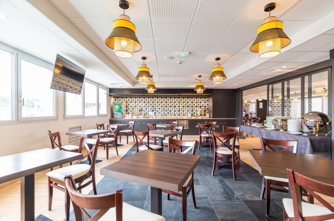 Salle de restaurant lumineuse avec tables en bois, chaises croisées et suspensions décoratives au plafond.