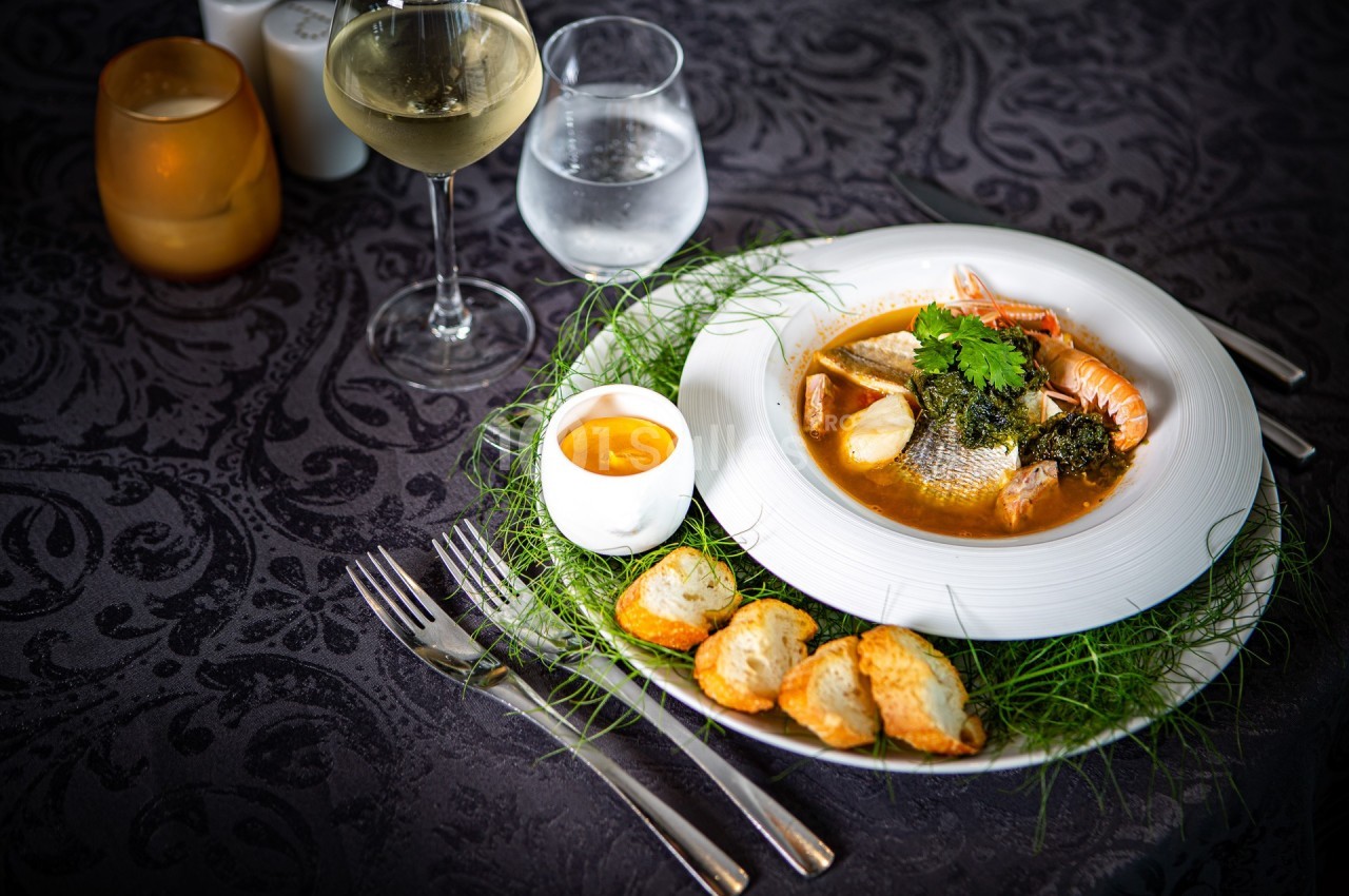 Assiette de bouillabaisse avec poissons, crustacés, croûtons, sauce et verres de vin et d'eau sur une table sombre.