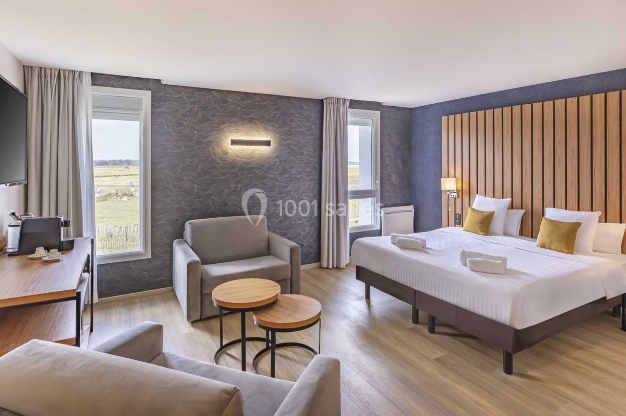 Chambre d'hôtel lumineuse avec lit double, coin salon, bureau, télévision murale et vue sur l'extérieur.