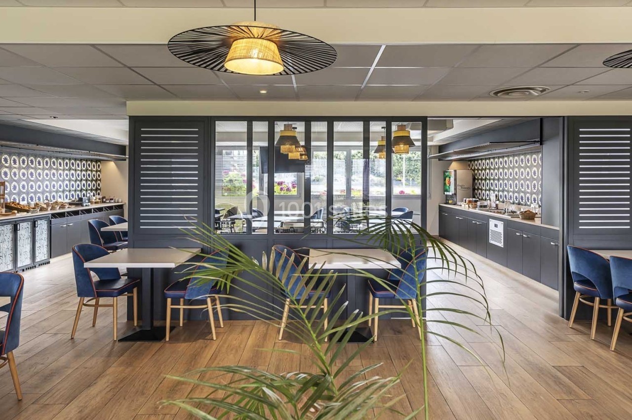 Salle de restaurant moderne avec tables, chaises bleues, luminaires suspendus et décoration murale géométrique.