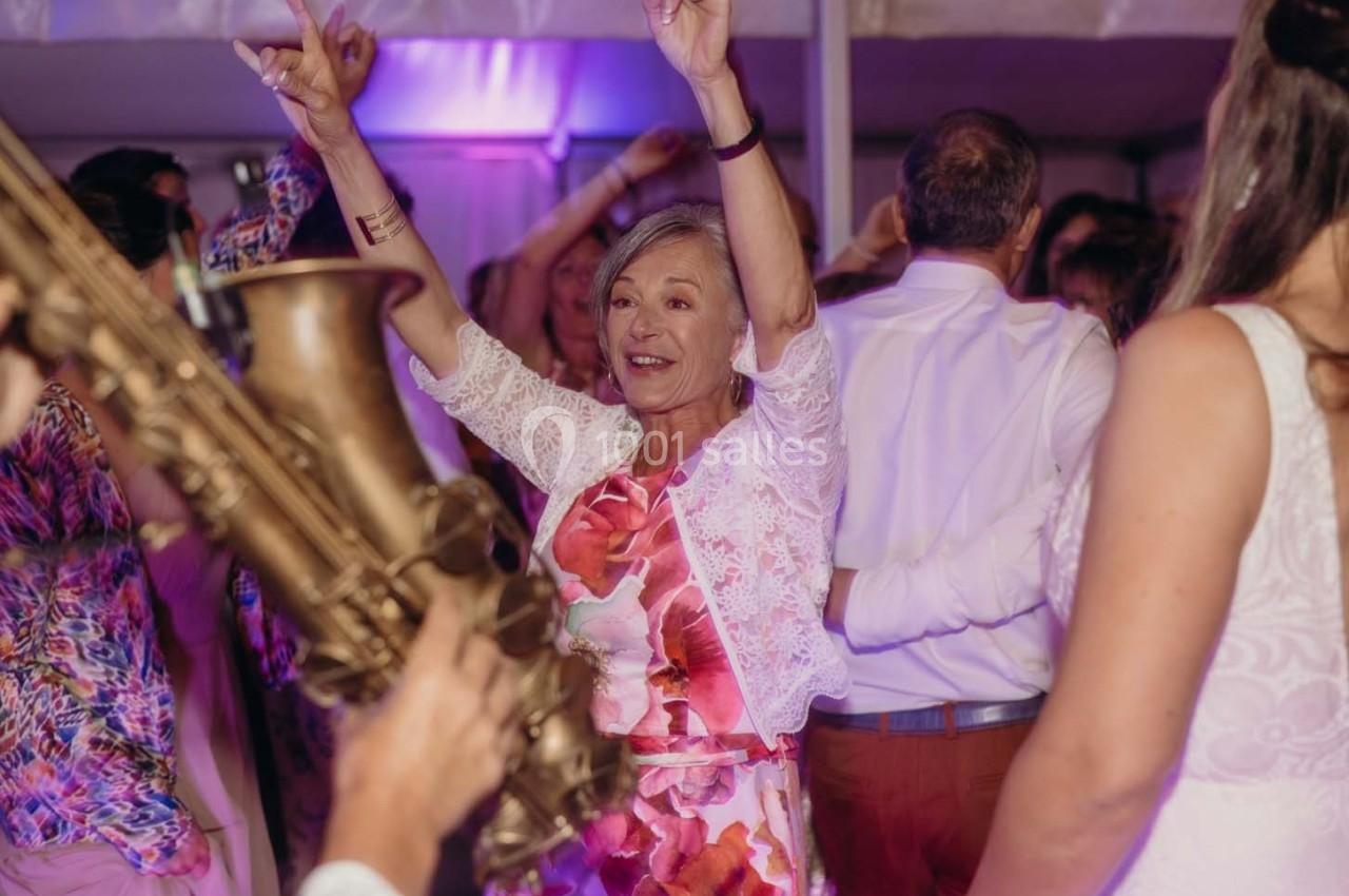 Une femme souriante danse lors d'une fête, entourée d'autres personnes et d'un saxophoniste au premier plan.