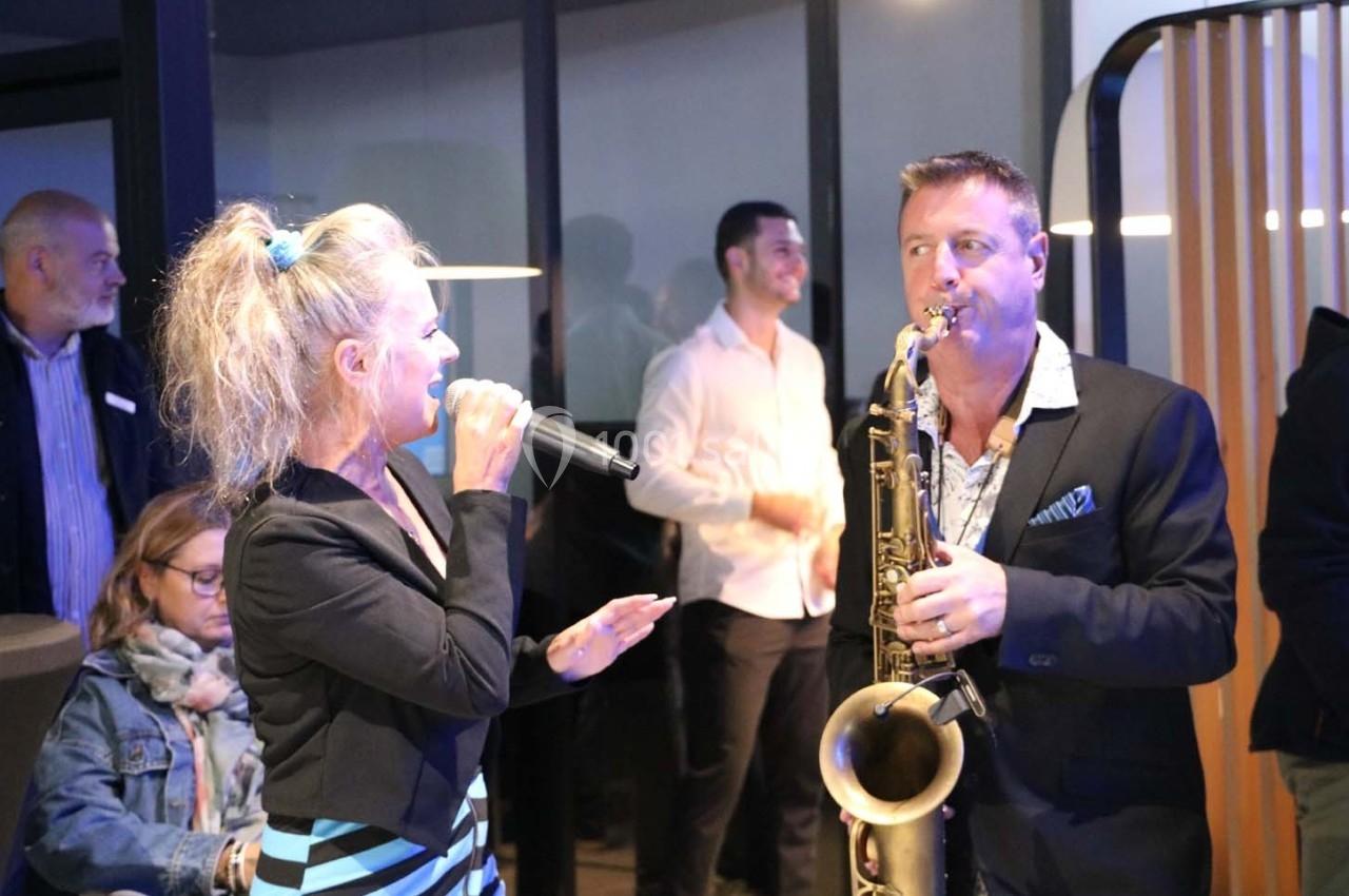 Une chanteuse et un saxophoniste jouent de la musique lors d'un événement en intérieur, entourés de spectateurs.
