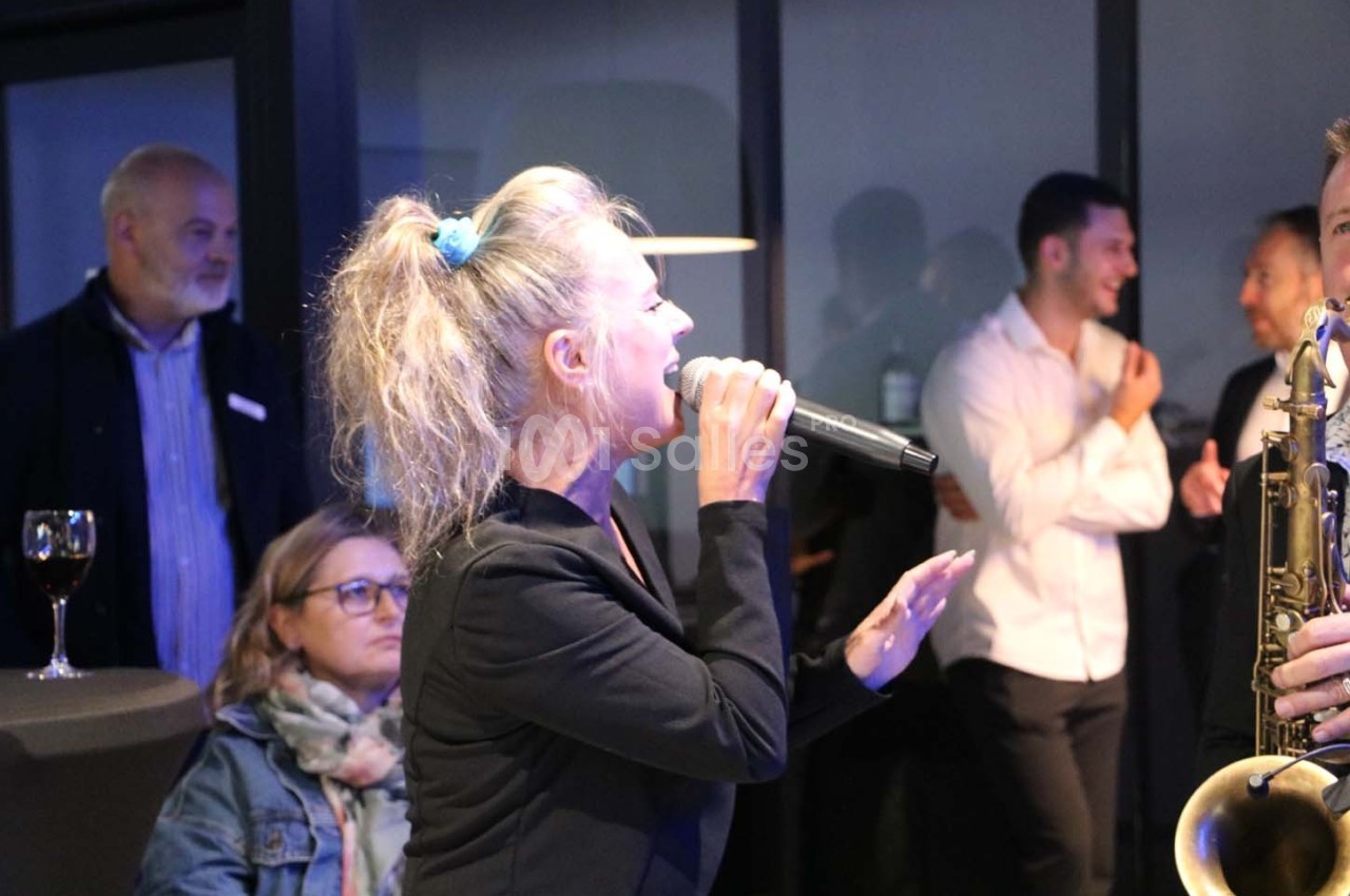 Une chanteuse interprète un morceau au micro lors d'un événement, entourée de spectateurs en arrière-plan.