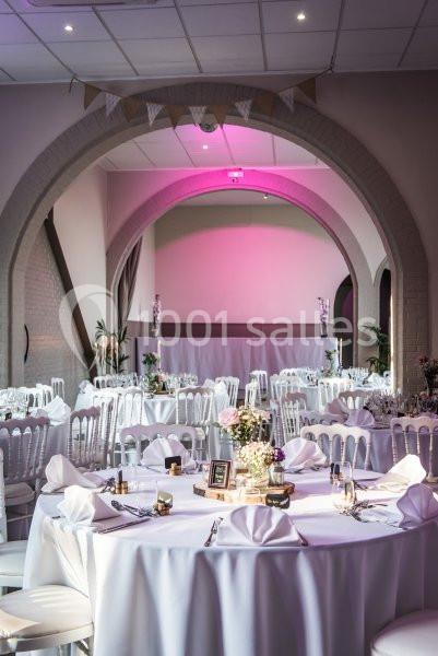 Salle de réception décorée avec des tables rondes, nappes blanches, chaises assorties et éclairage rose tamisé.