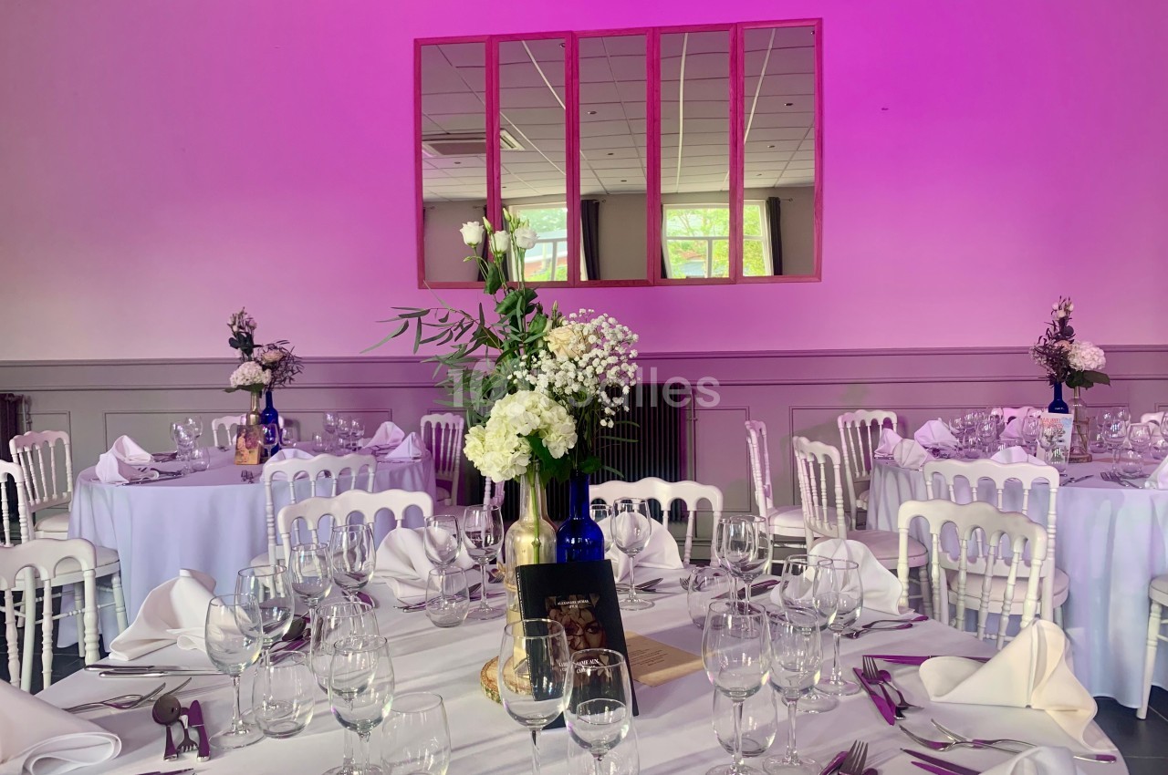 Salle de réception décorée avec des tables rondes, nappes blanches, verres, couverts et bouquets de fleurs.
