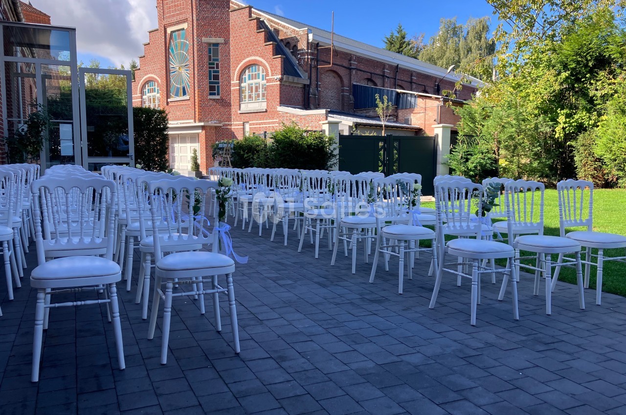 Chaises blanches alignées en extérieur sur une terrasse pavée, avec un bâtiment en briques et un jardin en arrière-plan.