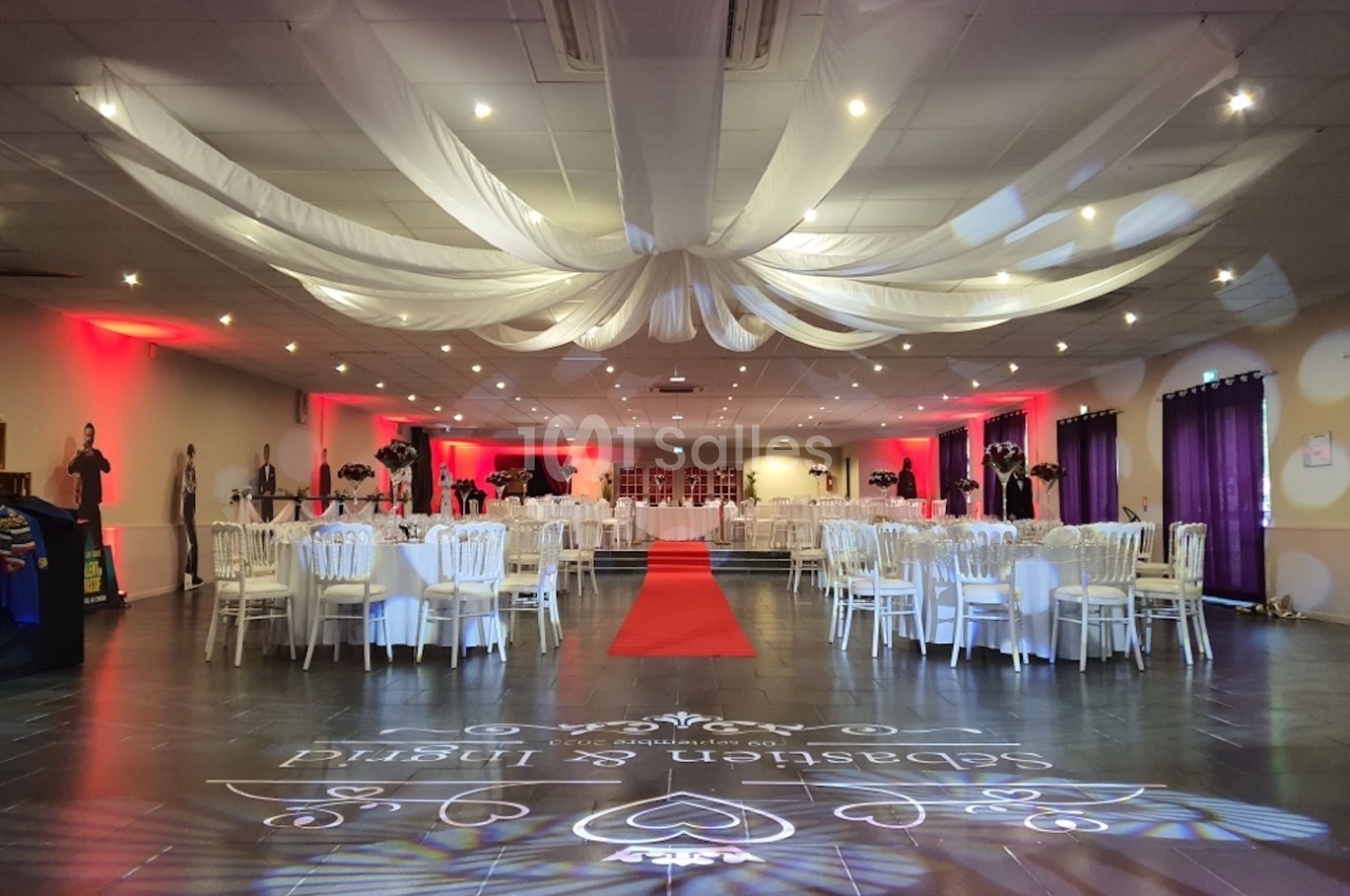 Salle de réception décorée avec des tables rondes, des chaises blanches, un tapis rouge et un plafond drapé de tissu blanc.