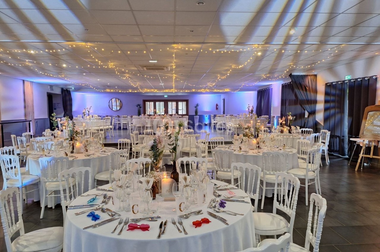 Salle de réception décorée avec des tables rondes dressées, chaises blanches et guirlandes lumineuses suspendues.