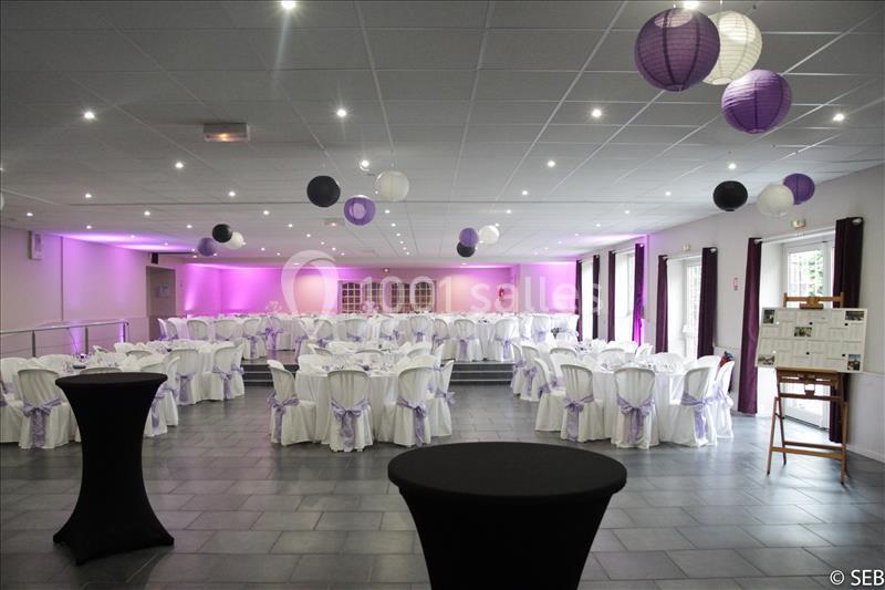 Salle de réception décorée avec des tables rondes, nappes blanches, nœuds violets et lanternes suspendues.