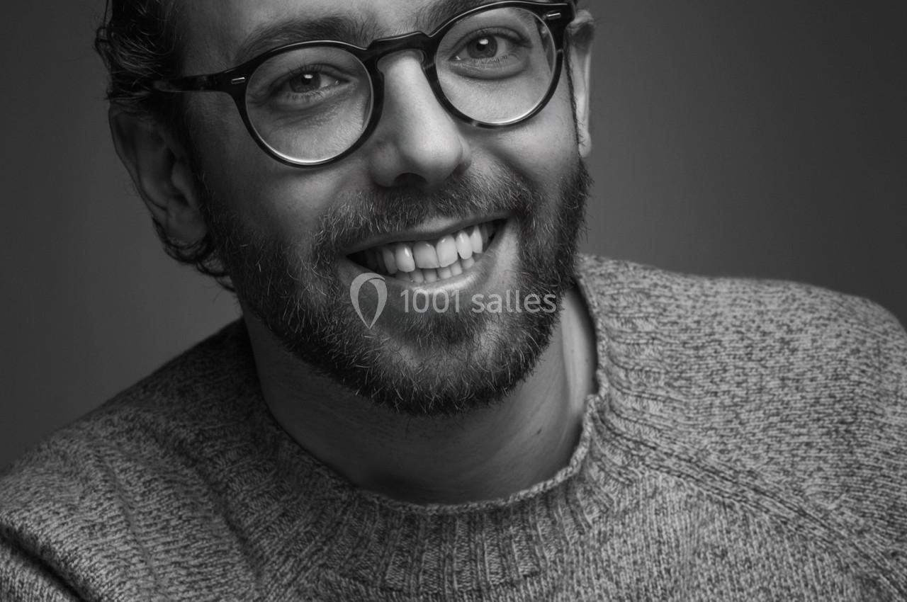 Portrait en noir et blanc d'un homme souriant portant des lunettes rondes et un pull en maille.
