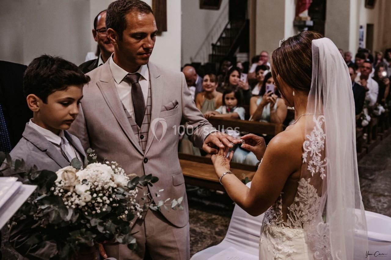 Une mariée passe une alliance au doigt du marié dans une église, entourés d'invités et d'un garçon d'honneur.
