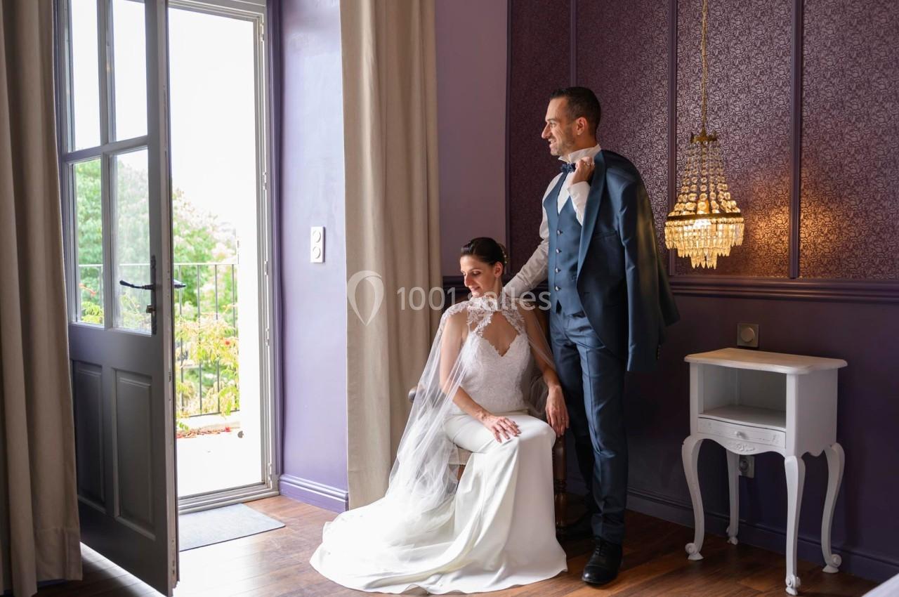 Un couple en tenue de mariage pose dans une pièce élégante avec un lustre et une vue sur un jardin par la porte ouverte.