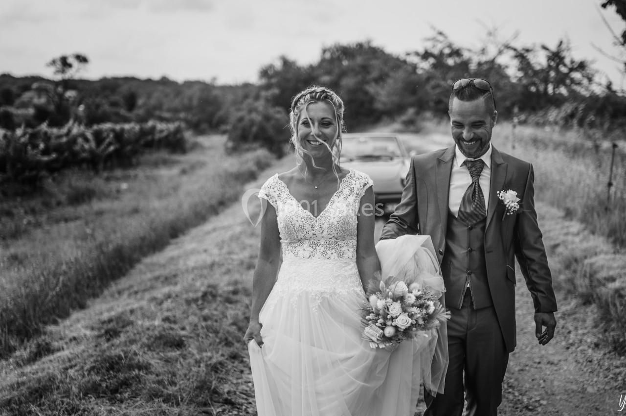 Un couple en tenue de mariage marche sur un chemin rural, souriant, avec des vignes en arrière-plan.