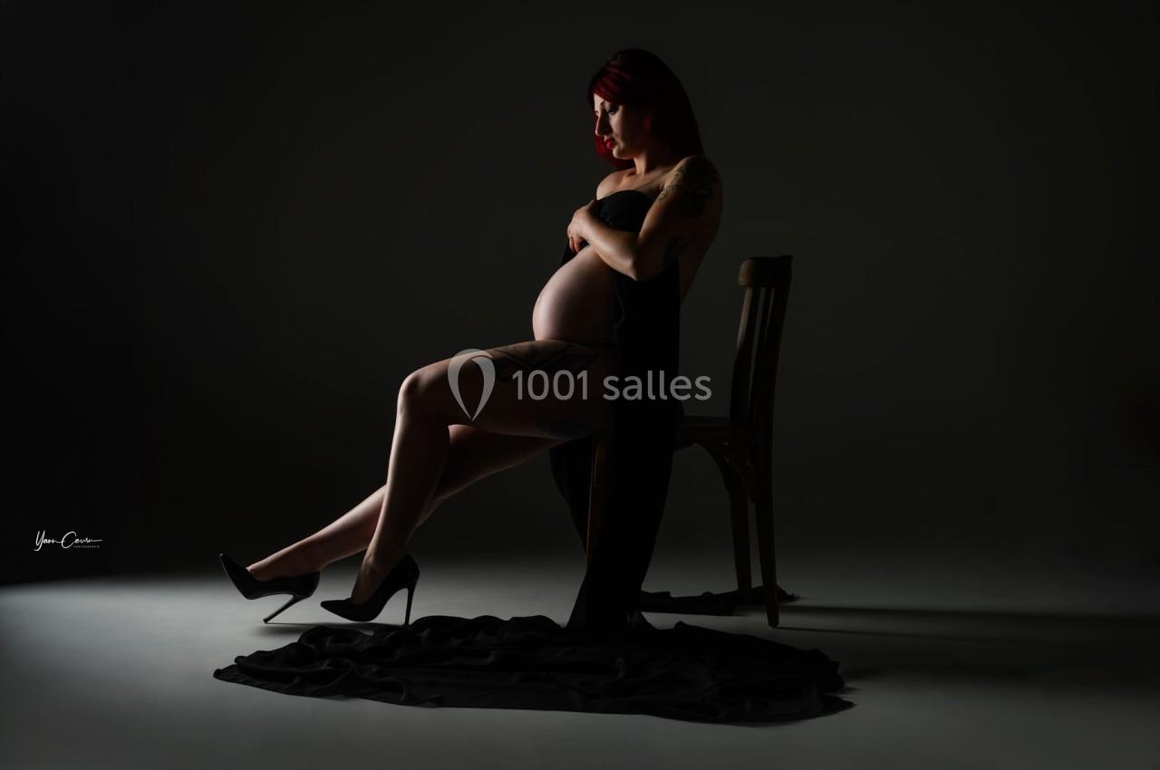 Femme enceinte assise sur une chaise dans un studio sombre, drapée d'un tissu noir, éclairée par une lumière douce.