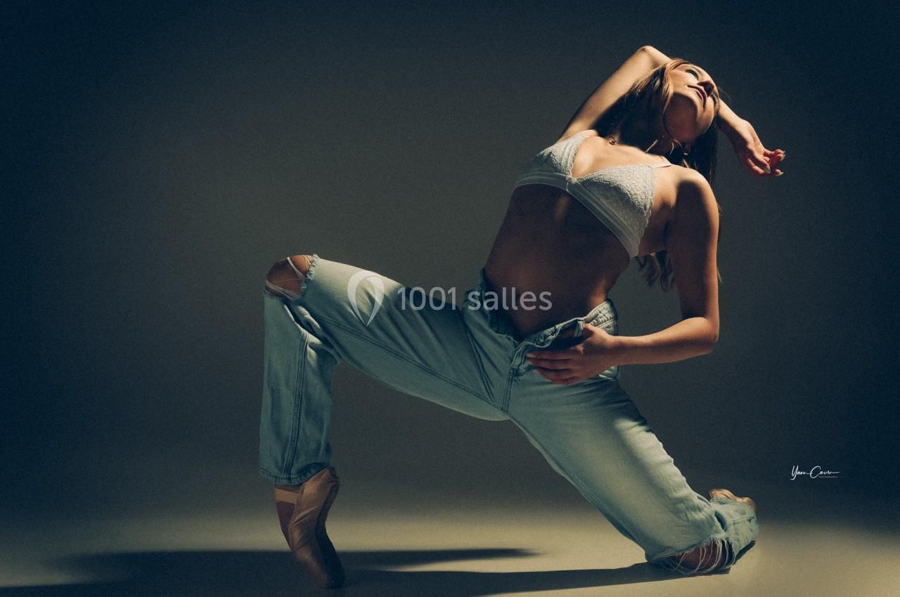 Une danseuse en jeans et brassière blanche effectue une pose artistique sur un sol éclairé dans une ambiance sombre.