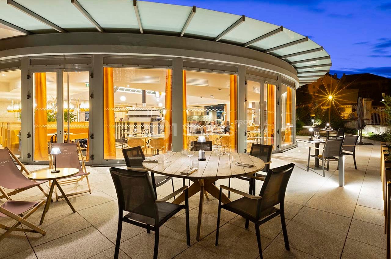 Terrasse d'un restaurant moderne avec tables et chaises, éclairée en soirée, vue sur l'intérieur lumineux à travers de…