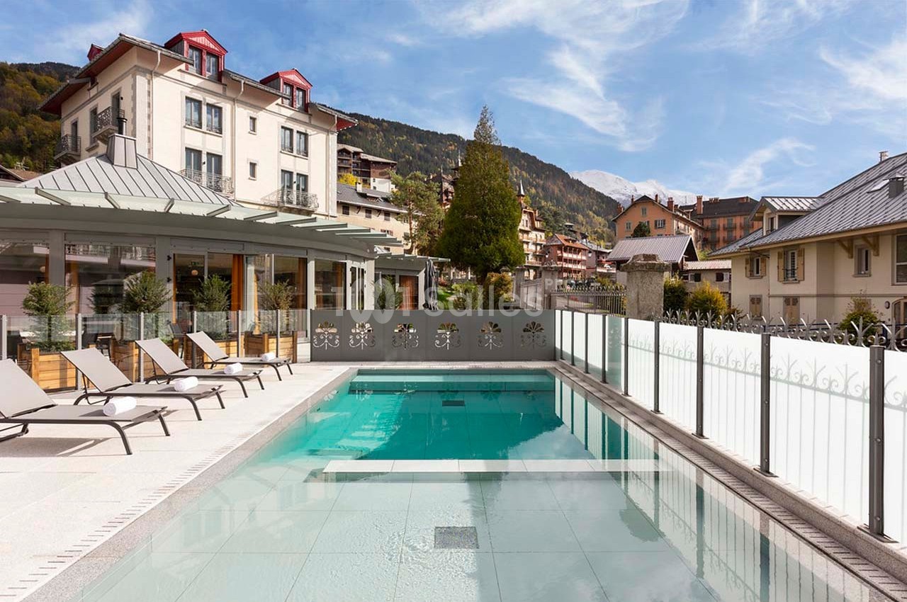 Piscine extérieure entourée de chaises longues, avec des bâtiments et des montagnes en arrière-plan par temps clair.