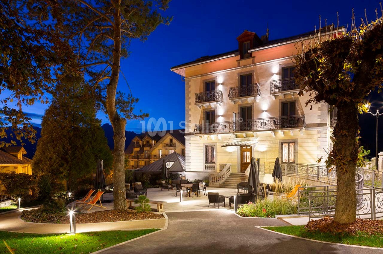 Façade d'un bâtiment éclairé la nuit, entouré d'un jardin avec des arbres, des chaises et des tables en extérieur.