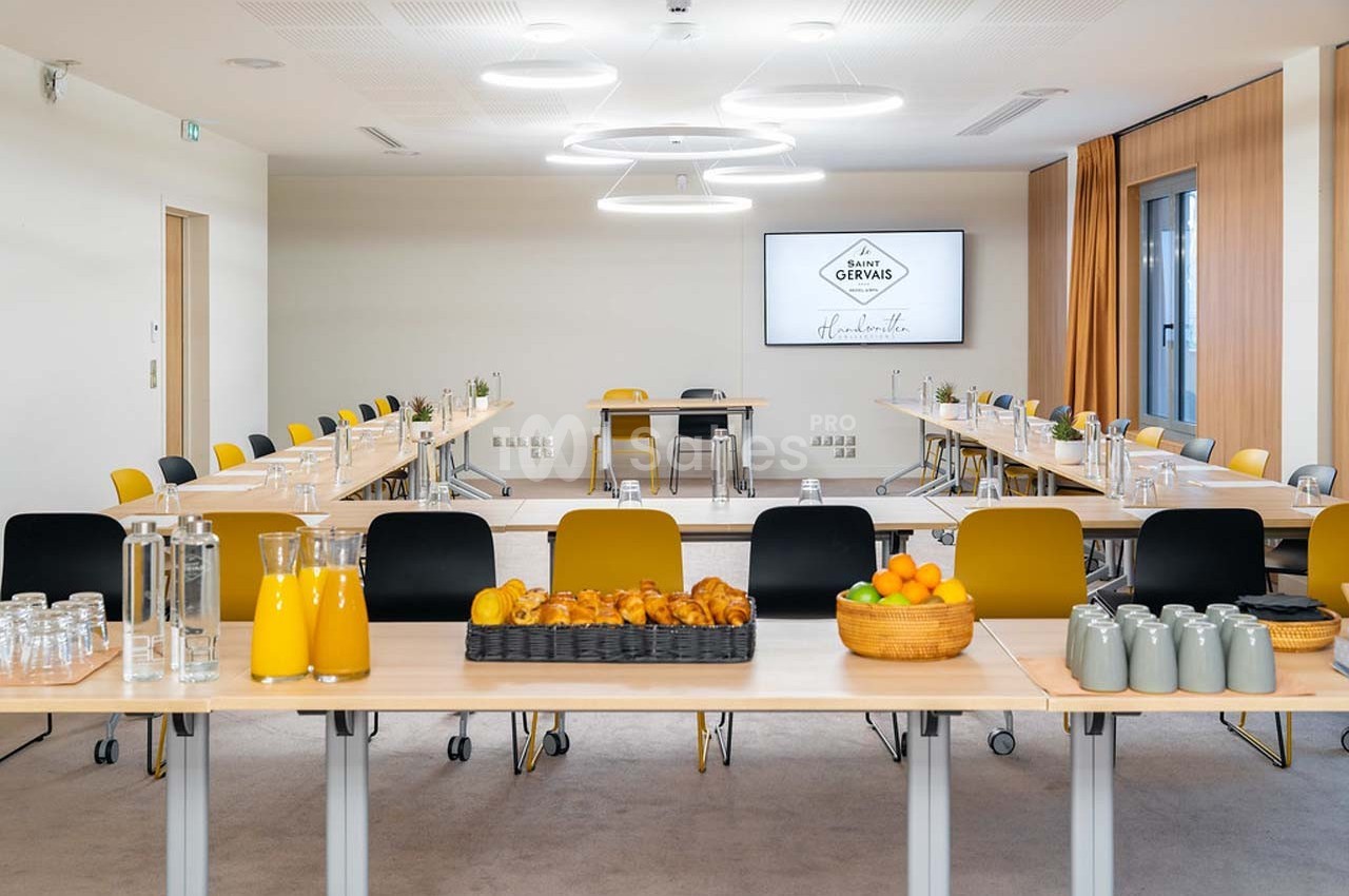 Salle de réunion lumineuse avec tables disposées en U, boissons, viennoiseries et fruits sur une table centrale.