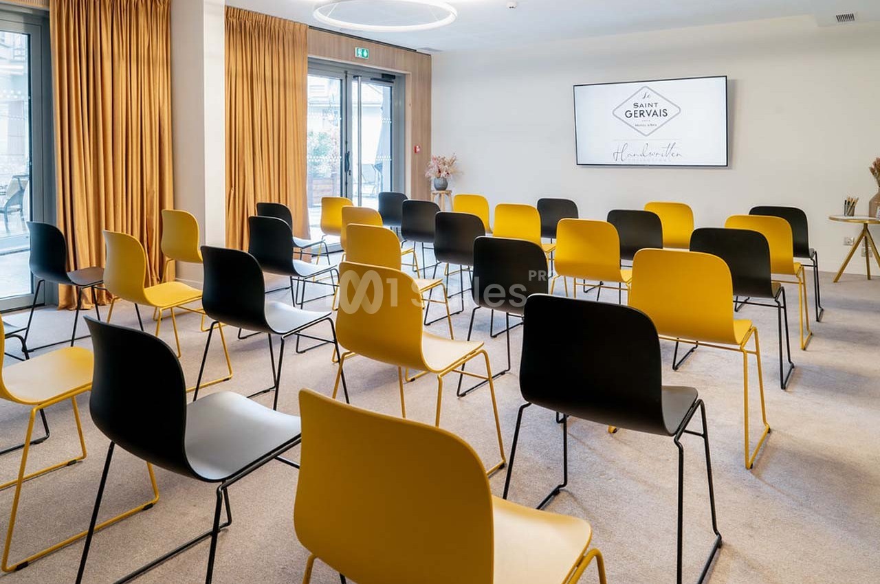 Salle de réunion lumineuse avec des chaises noires et jaunes disposées face à un écran mural.