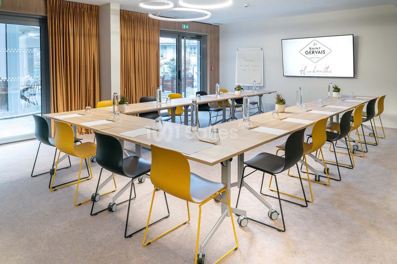 Salle de réunion lumineuse avec tables disposées en U, chaises colorées, écran, tableau blanc et rideaux dorés.