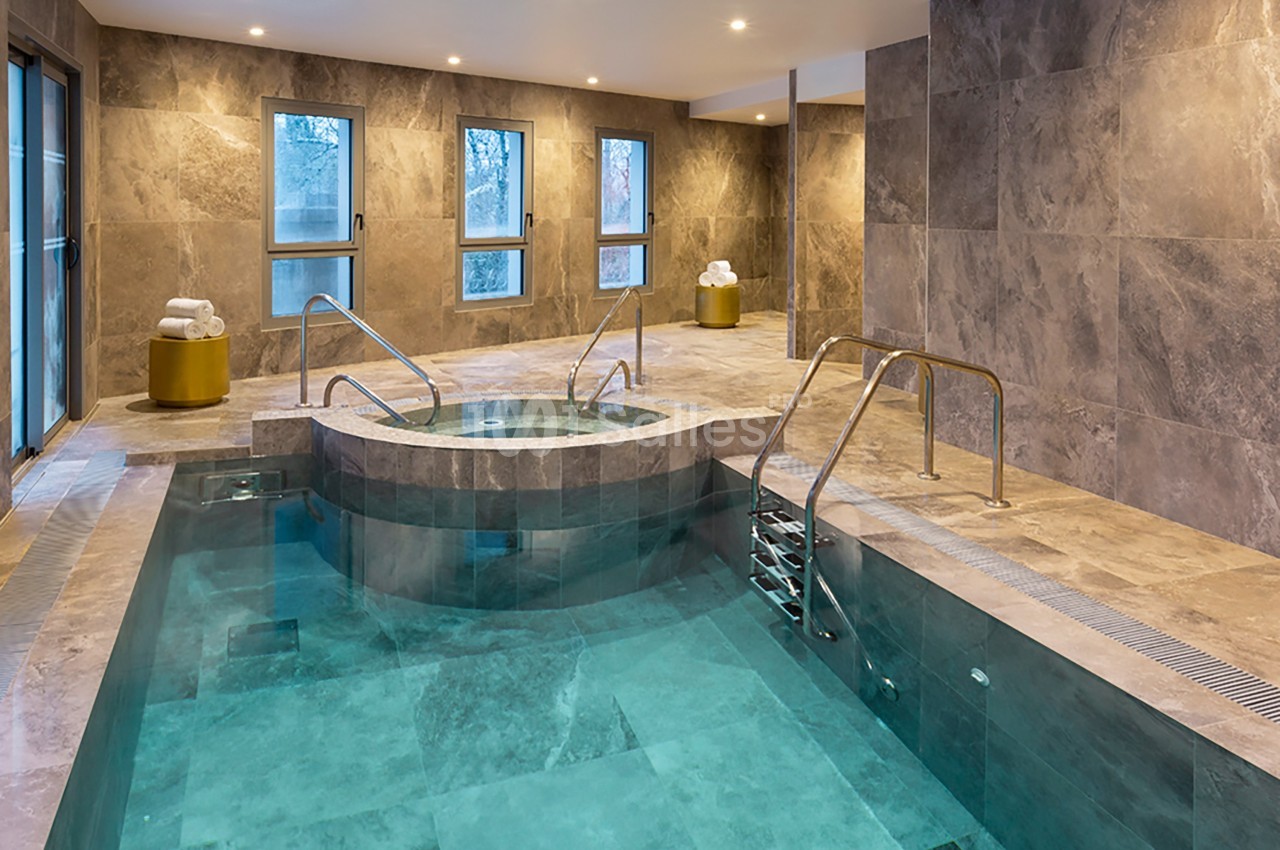 Piscine intérieure avec jacuzzi intégré, entourée de murs en pierre grise et éclairée par des fenêtres et des spots au…