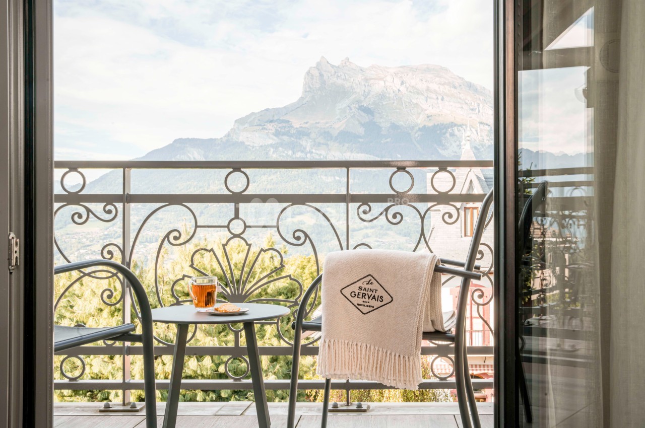 Vue d'un balcon avec table, tasse de thé et plaid, donnant sur une montagne sous un ciel partiellement nuageux.