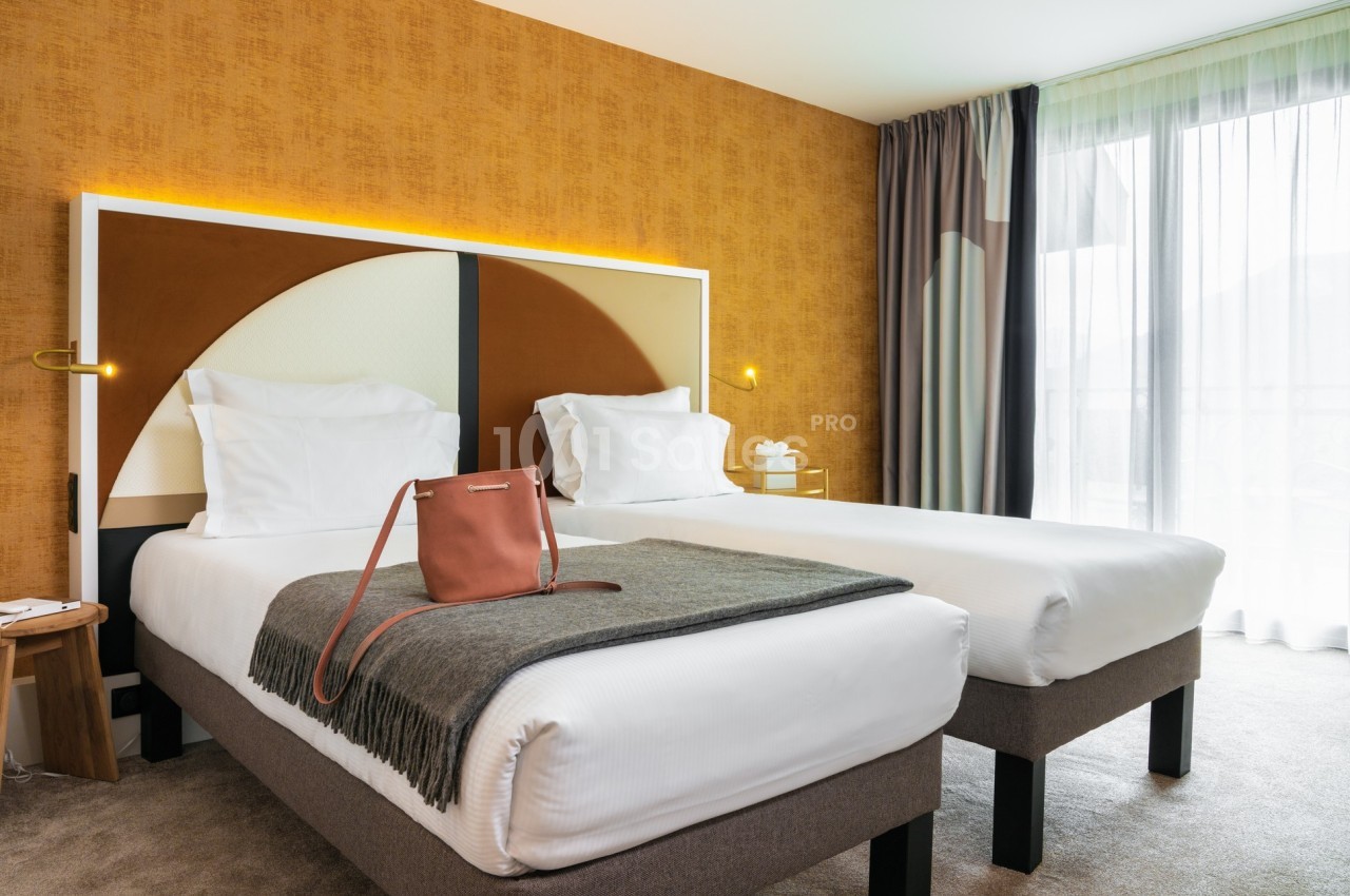 Deux lits simples avec tête de lit moderne, plaid gris et sac posé, dans une chambre lumineuse aux tons chauds.