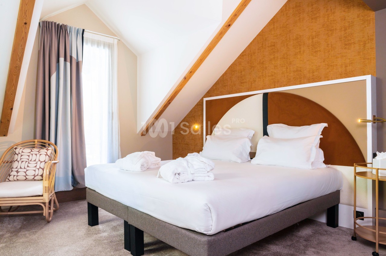 Chambre lumineuse sous combles avec lit double, fauteuil en rotin, rideaux épais et décoration murale moderne.