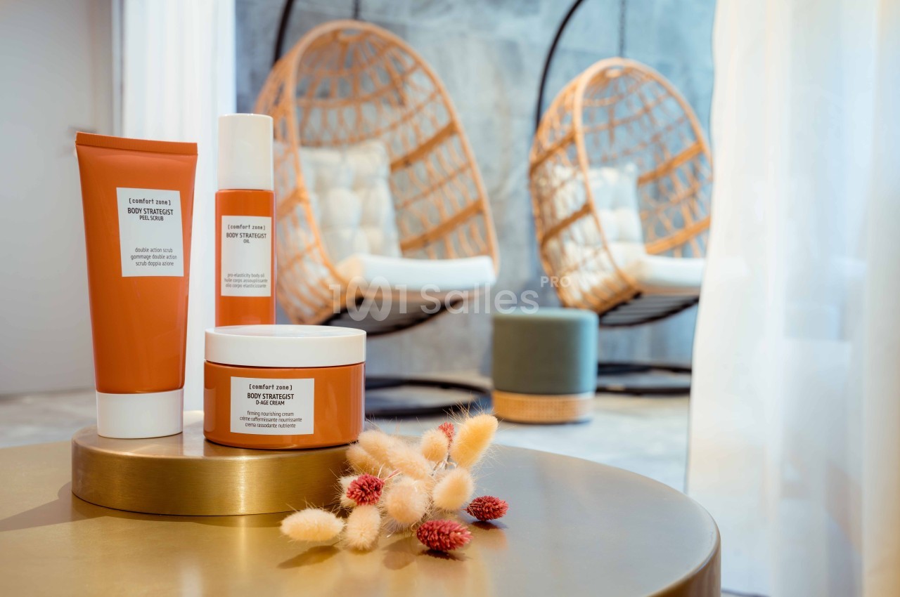 Produits cosmétiques sur une table dorée avec des fauteuils suspendus et des fleurs séchées en arrière-plan.