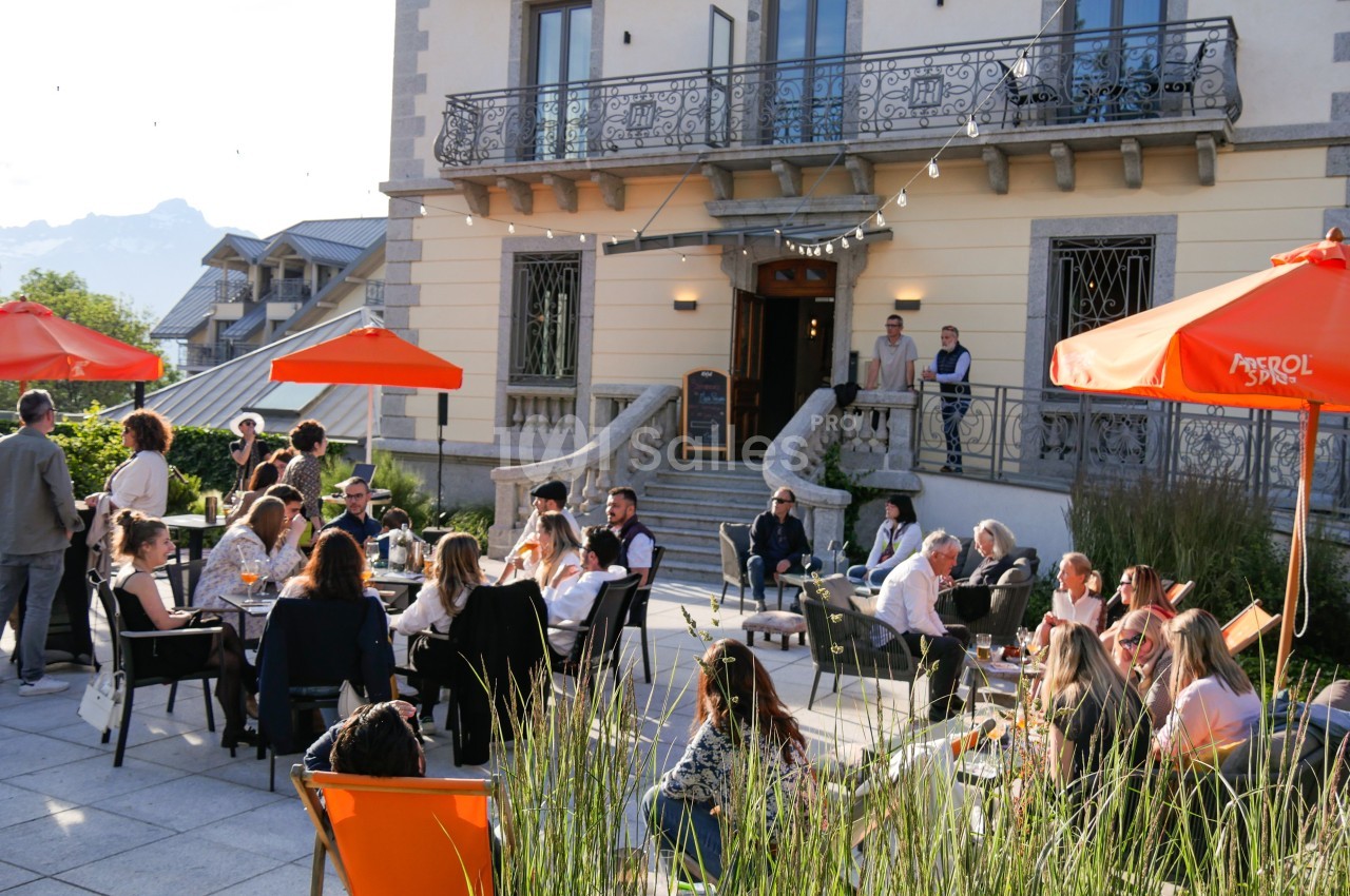 Groupe de personnes assises et debout sur une terrasse ensoleillée devant un bâtiment avec des parasols orange.
