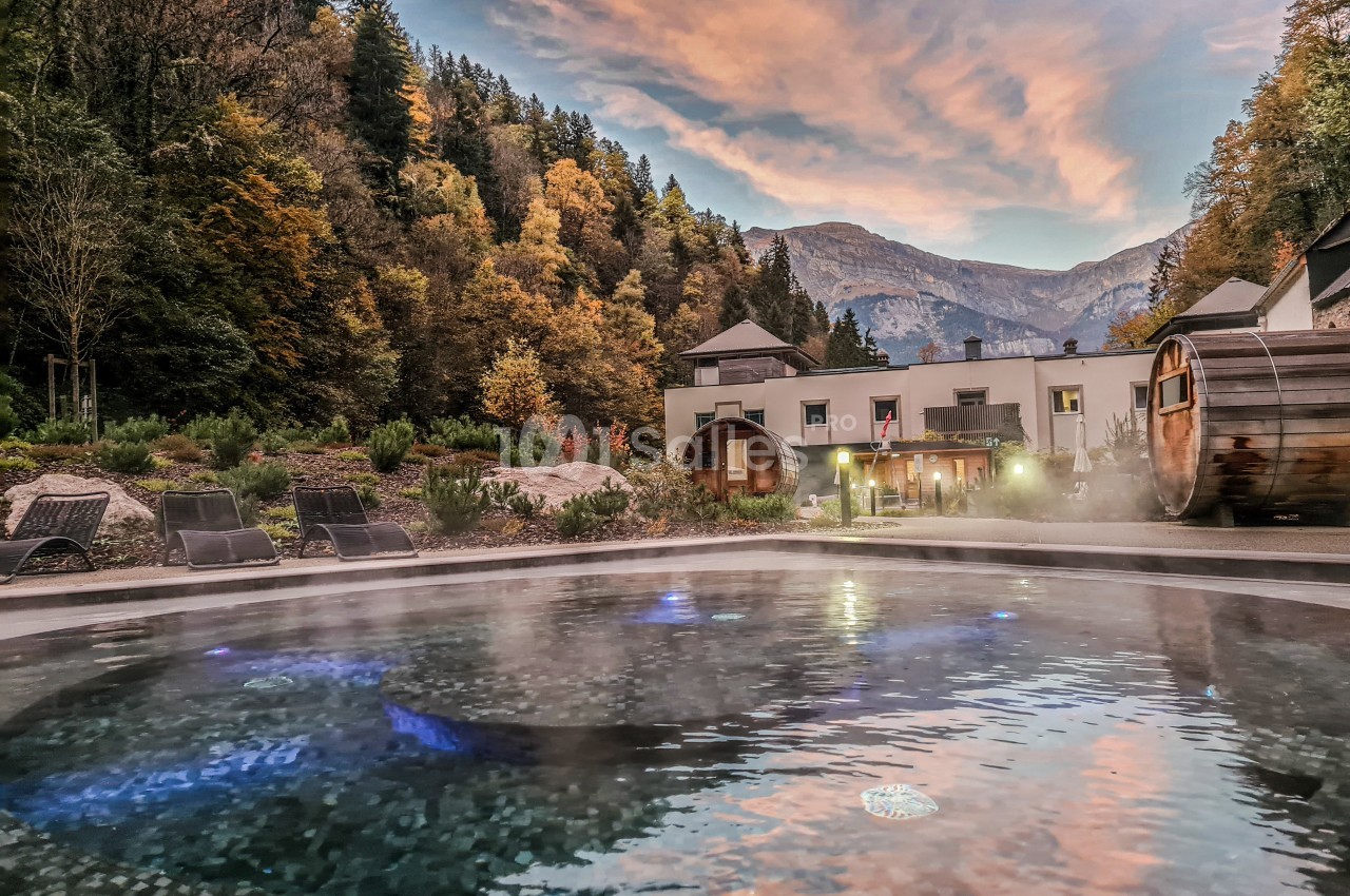 Vue d'un bassin extérieur fumant entouré de chaises longues, avec des montagnes et une forêt en arrière-plan au coucher du…