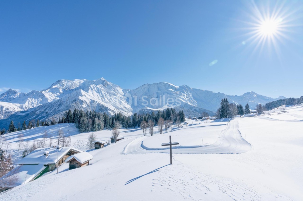 Paysage enneigé avec montagnes en arrière-plan, chalets dispersés et ciel dégagé sous un soleil éclatant.