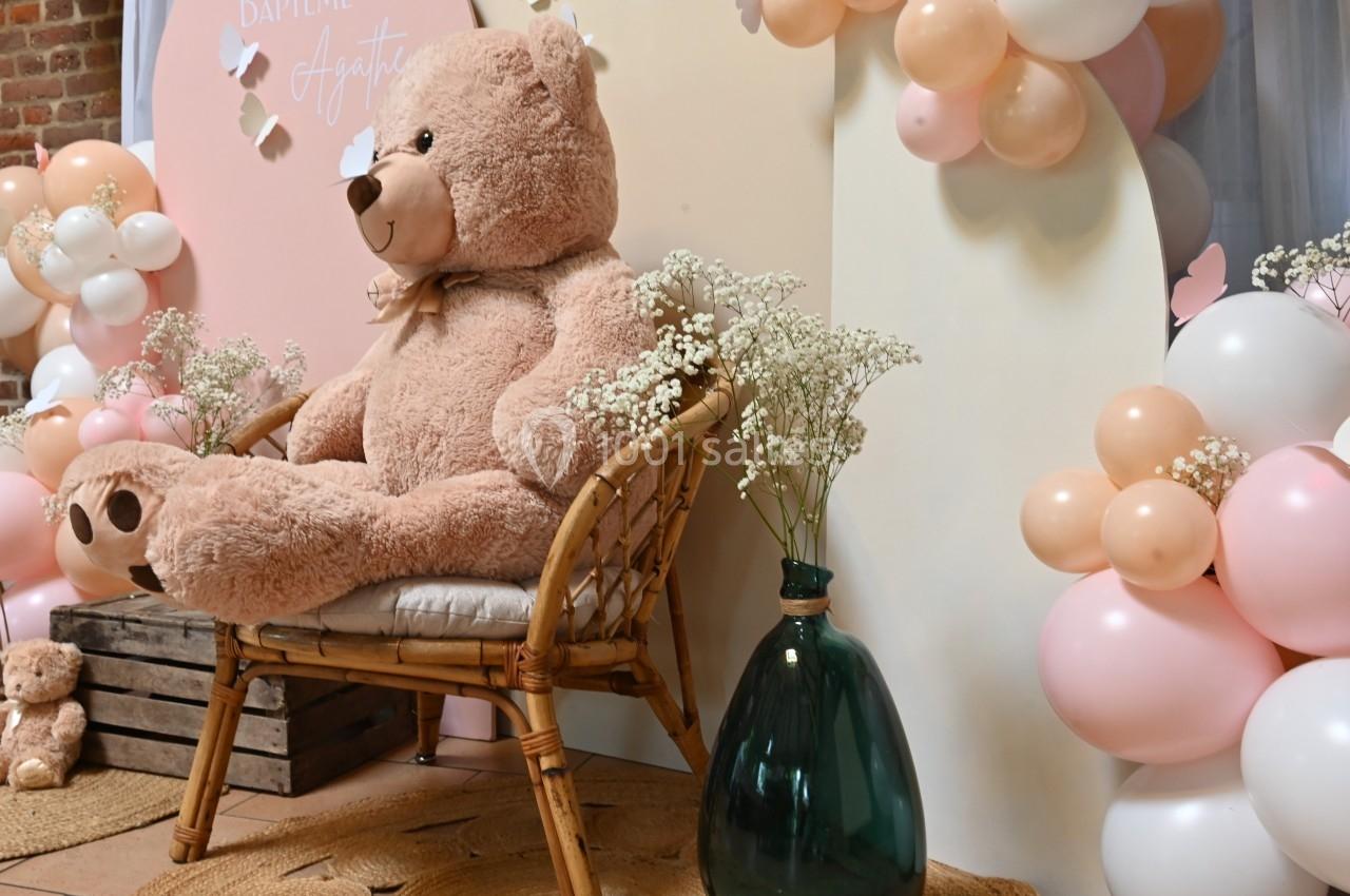 Un grand ours en peluche beige est assis sur une chaise en rotin, entouré de ballons roses et blancs et de fleurs.