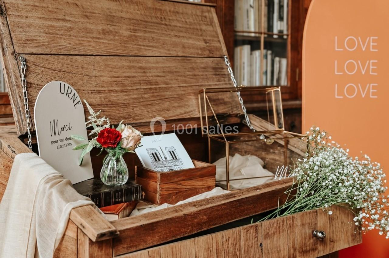 Coffre en bois ouvert contenant des fleurs, une urne, des livres et des objets décoratifs, avec une affiche en arrière-plan.