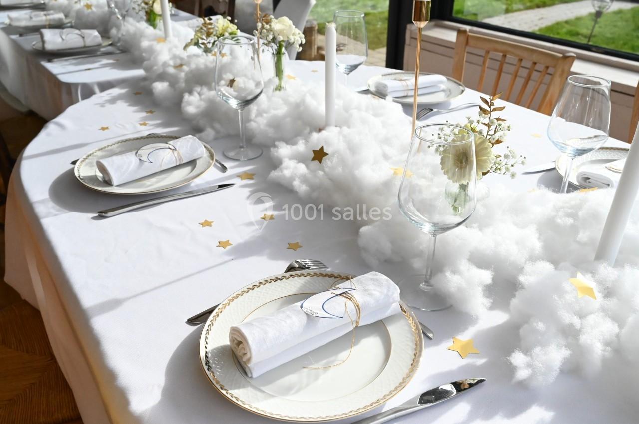 Table décorée avec une nappe blanche, des assiettes élégantes, des bougies, des fleurs et des éléments évoquant des nuages.