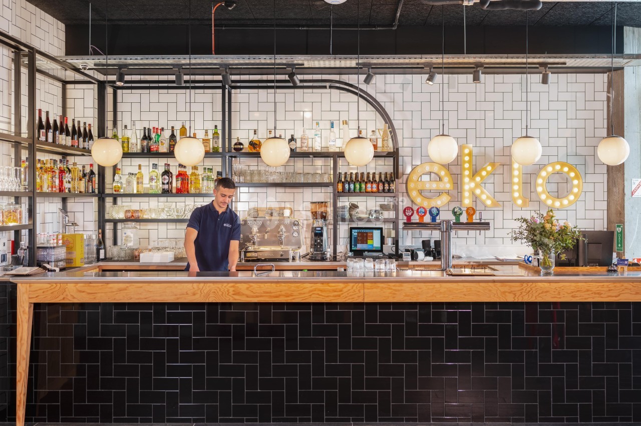 Bar moderne avec comptoir en bois, carrelage mural blanc, étagères garnies de bouteilles et lumières suspendues.