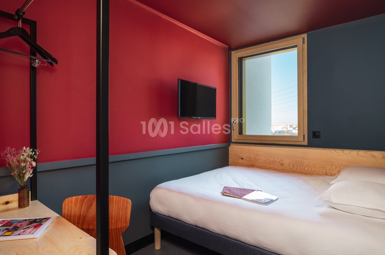 Chambre avec lit double, bureau, chaise, télévision murale, fenêtre et décoration aux tons rouges et bois.