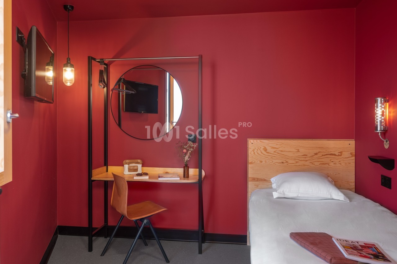 Chambre avec murs rouges, lit simple, bureau en bois avec miroir rond, chaise et éclairage suspendu.