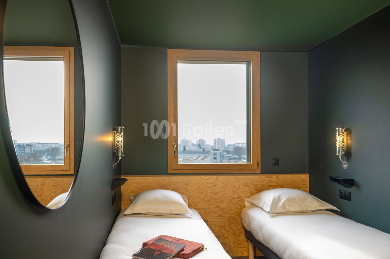 Deux lits simples avec linge blanc dans une chambre moderne, éclairée par deux fenêtres avec vue sur la ville.