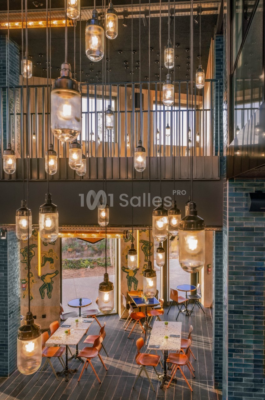 Suspensions lumineuses en verre éclairant un espace intérieur avec tables, chaises orange et murs en briques bleues.
