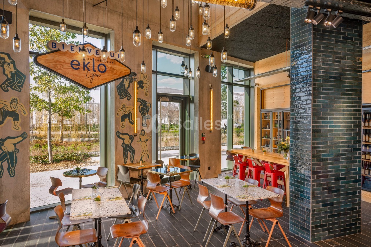 Salle de restaurant lumineuse avec tables en bois, chaises variées, murs décorés et grandes baies vitrées donnant sur l…