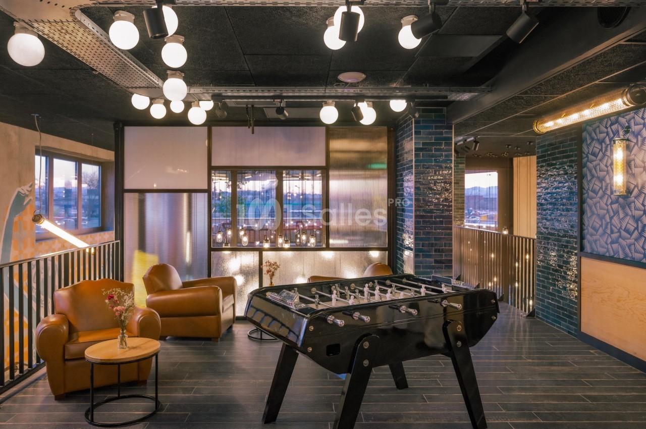 Salle de détente moderne avec baby-foot, fauteuils en cuir, éclairage suspendu et décoration contemporaine.