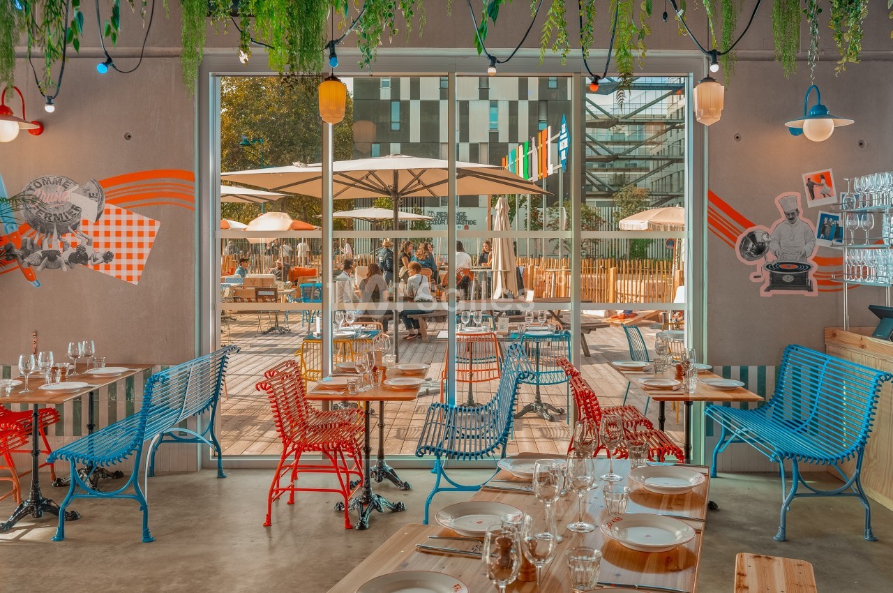 Salle de restaurant colorée avec des tables dressées, donnant sur une terrasse ensoleillée avec des parasols.