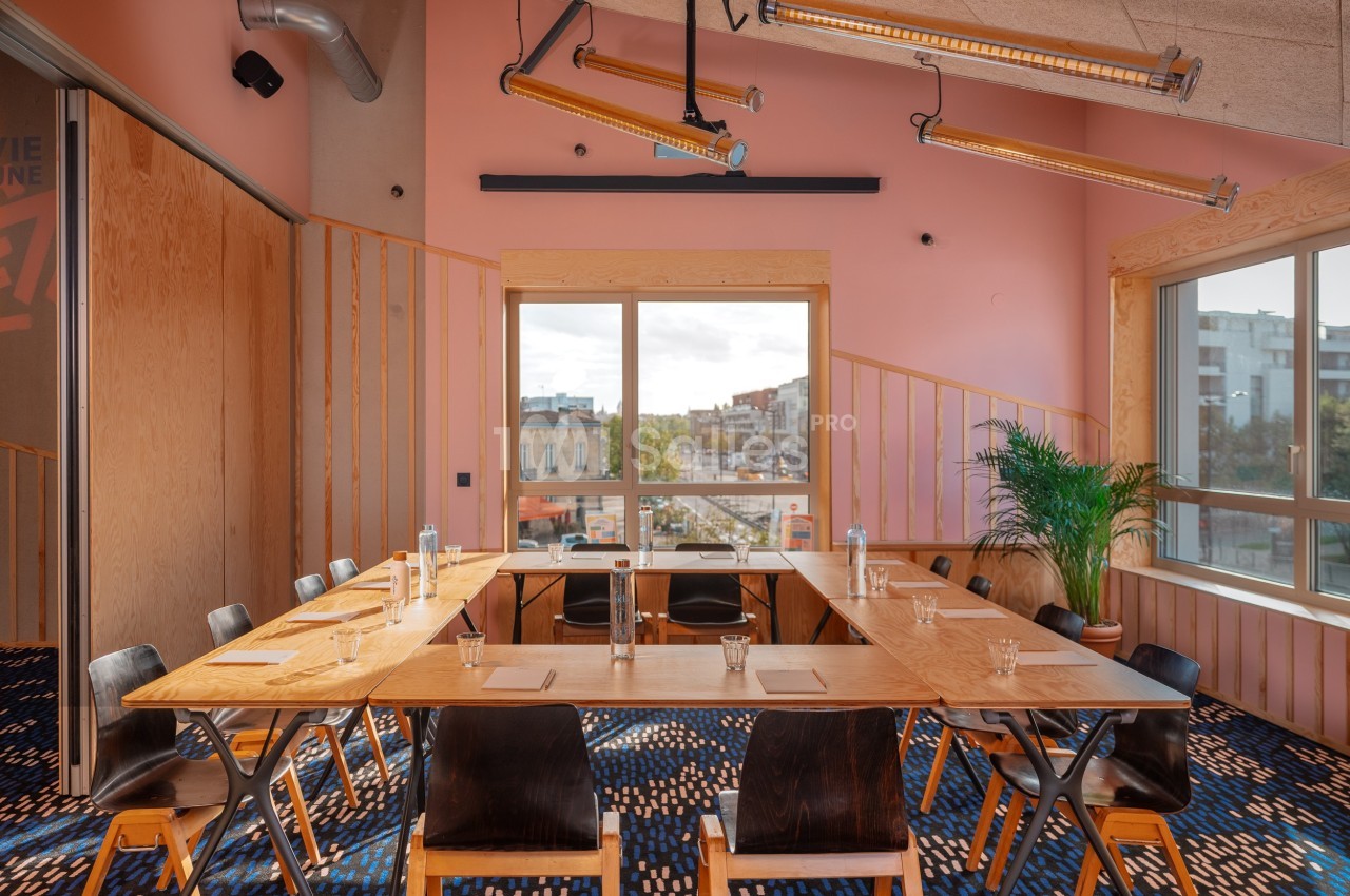 Salle de réunion lumineuse avec grande table en bois, chaises noires, plantes et vue sur l'extérieur par une large fenêtre.