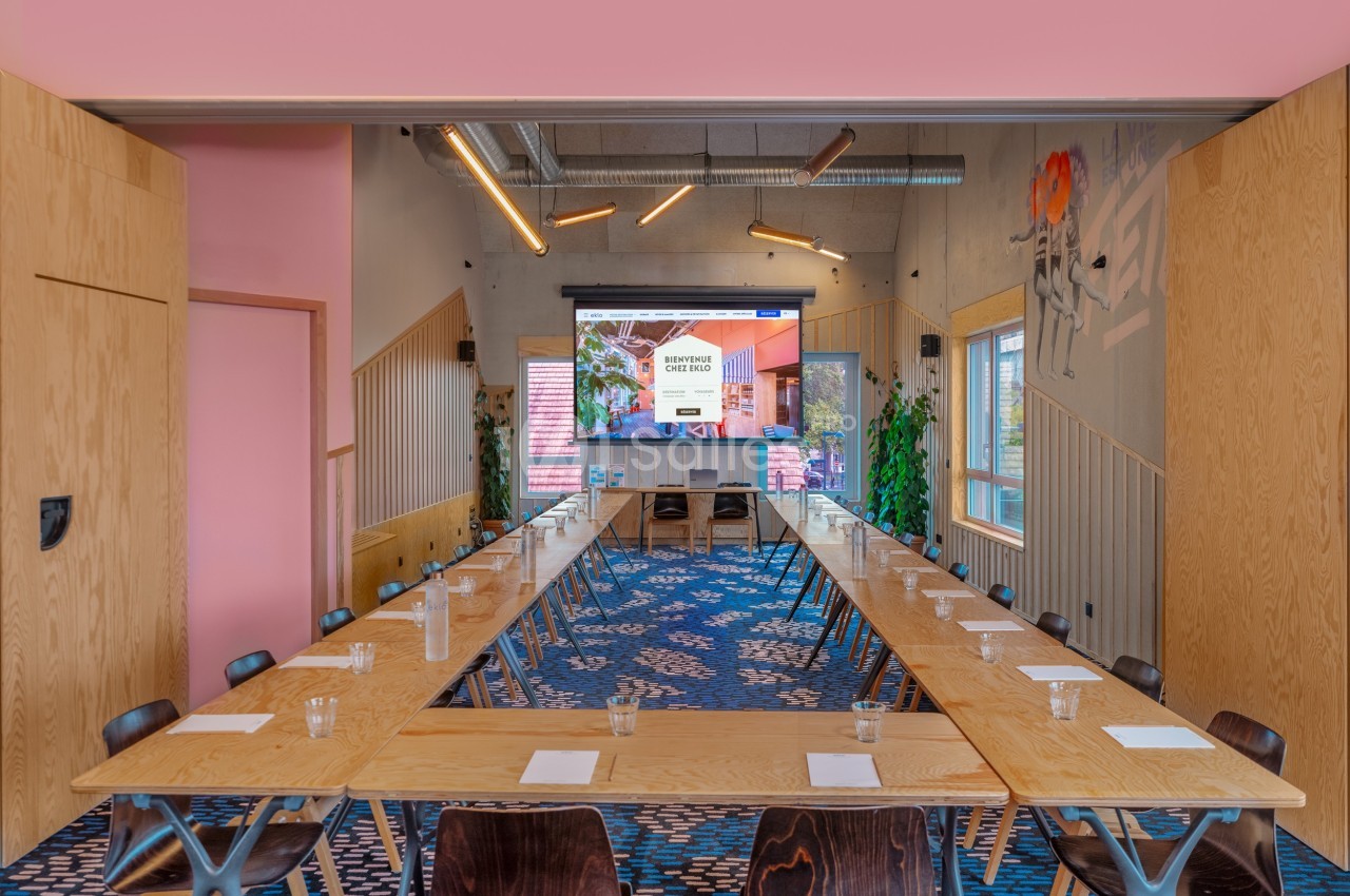 Salle de réunion moderne avec tables en U, chaises en bois, écran de présentation et décorations murales colorées.