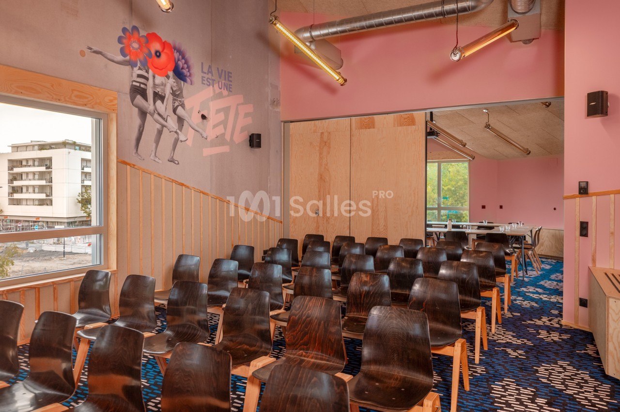 Salle de réunion colorée avec des chaises en bois, mur rose décoré d'une fresque et grande fenêtre donnant sur l'extérieur.